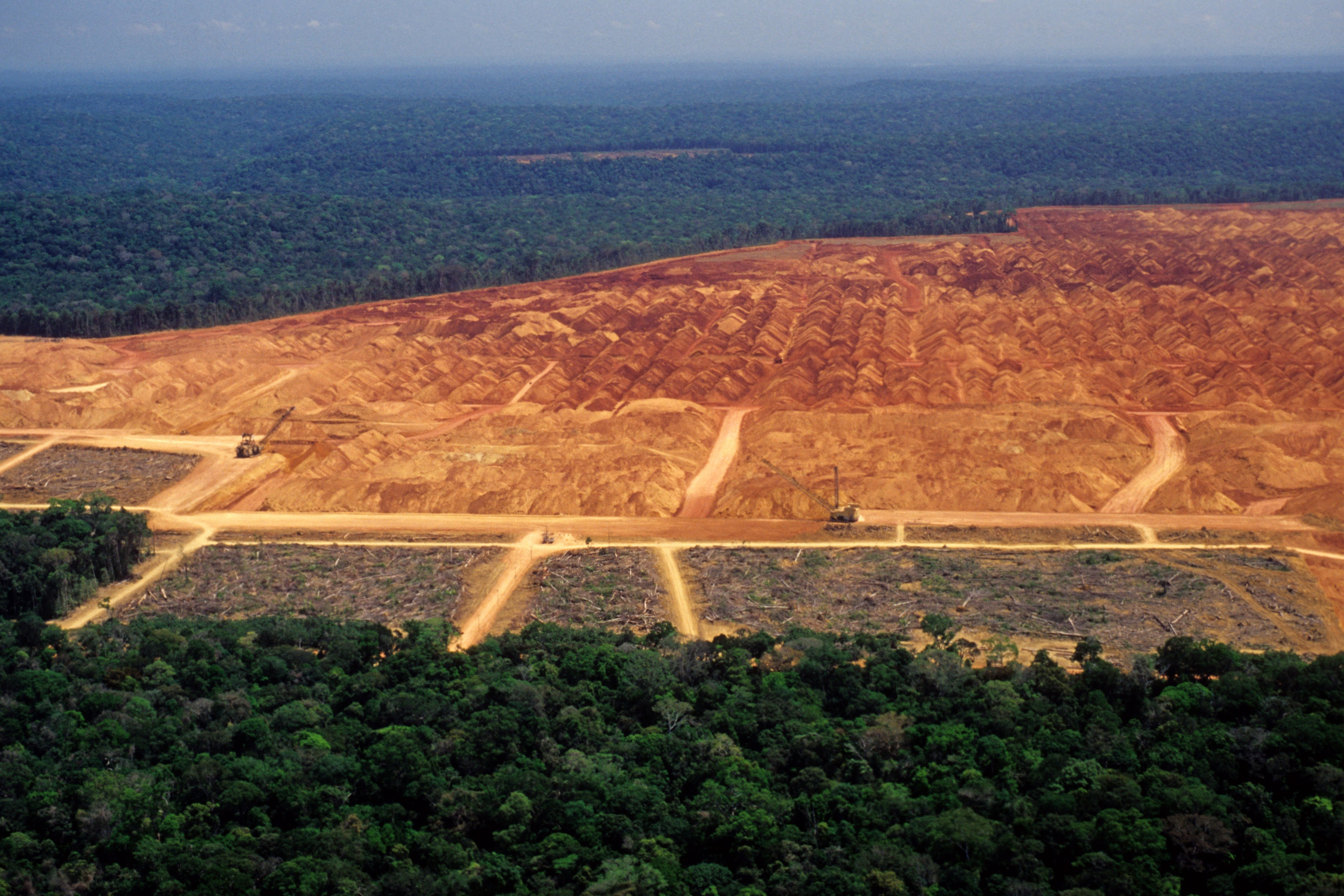 Deforestation in the amazon with red soil exposed with intact forest surrounding the area. Heavy machinery can be seen in the area.