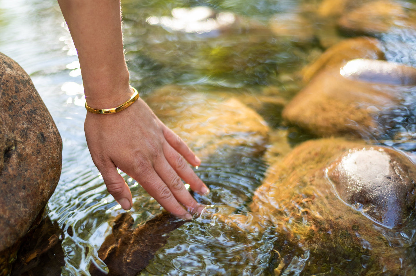 A gold counterbalance bracelet is worn by a model who dips their hand into the water of the rattlesnake creek.
