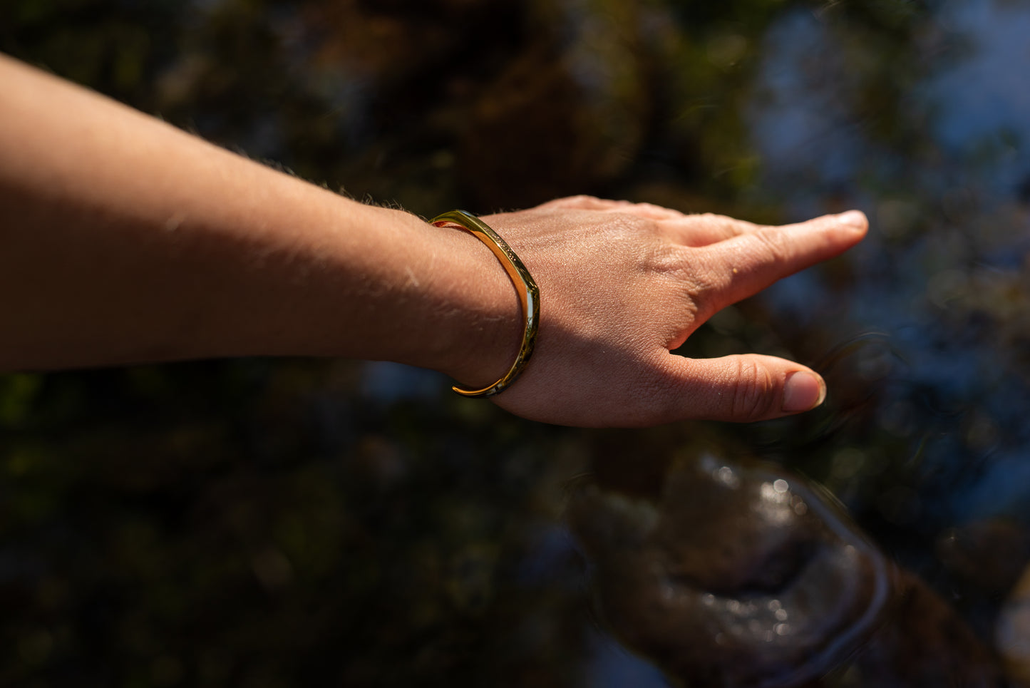 A model wearing the yellow gold counterbalance bracelet reaches towards a reflect pool of a natural creek.