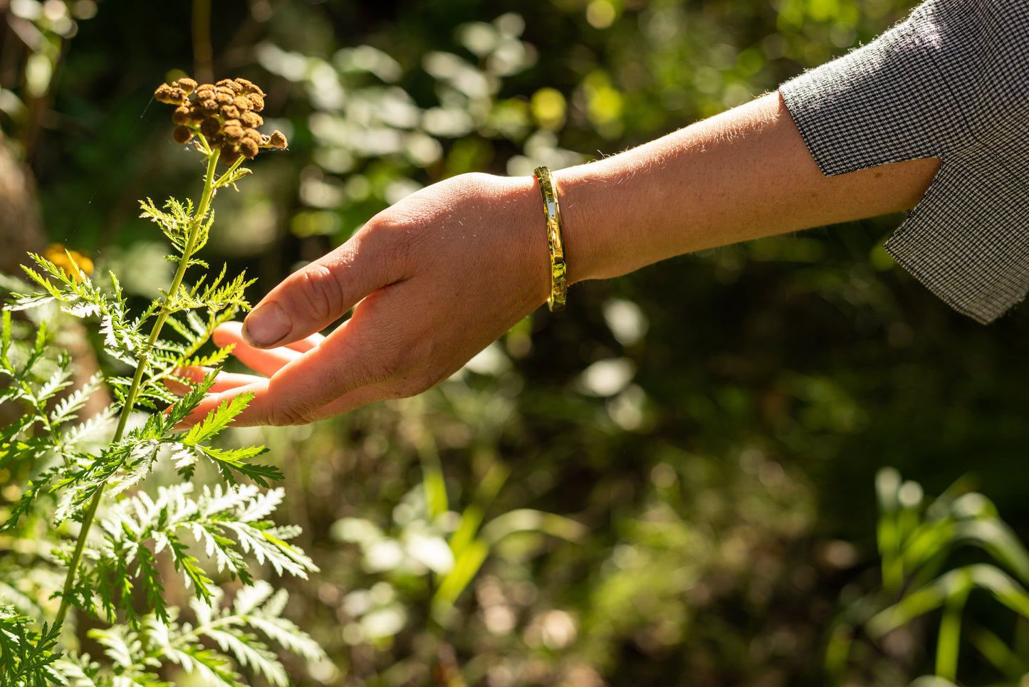 A gold counterbalance bracelet engraved with the abbreviations for 30 tons of carbon dioxide equivalent. The bracelet is worn by a model who reaches out to a plant with a green forest in the background.