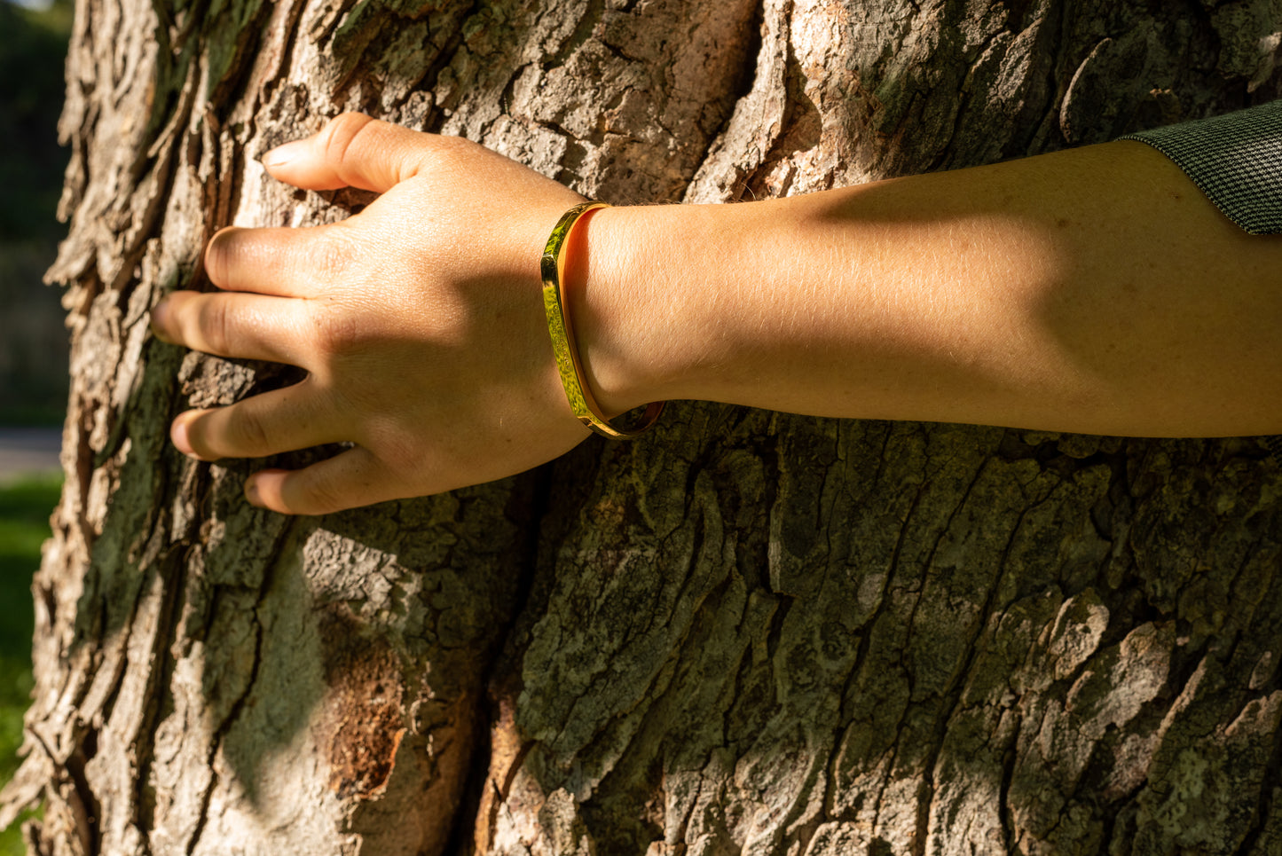 A model wearing the yellow gold counterbalance bracelet gestures wraps an arm around the trunk of a tree. The forest floor is reflected onto the bracelet with vibrant green color.