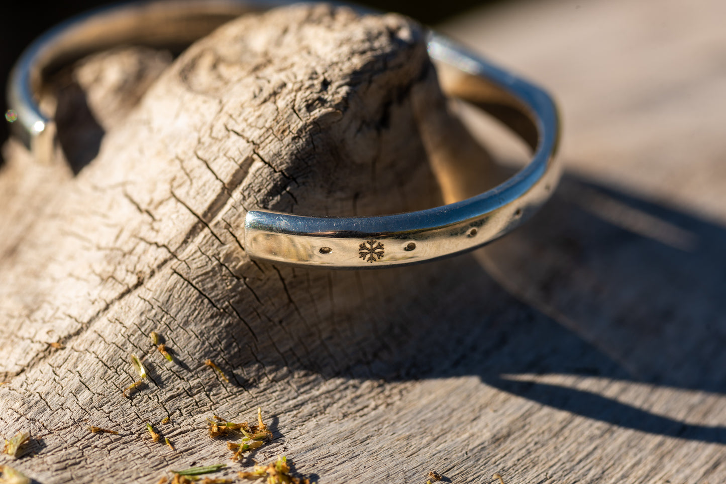 A silver counterbalance bracelet is displayed on a piece of smooth driftwood with a refrigerant management snowflake shaped charm engraved onto the right side.