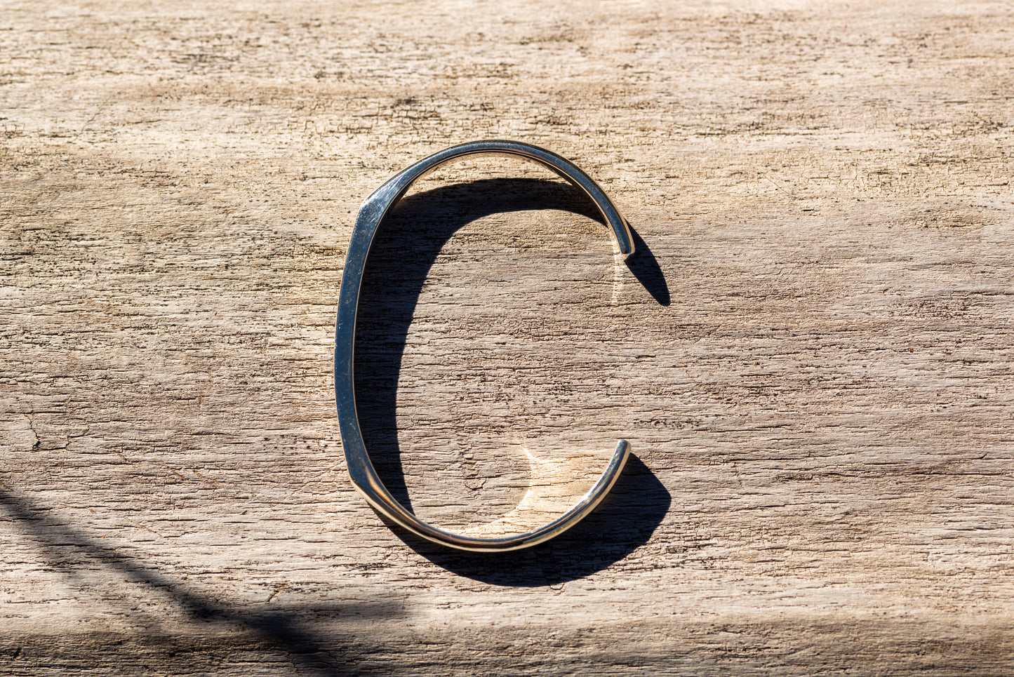 A silver counterbalance bracelet laying on smooth driftwood in the sunshine, with light reflecting off of it and thin shadows cast behind it.