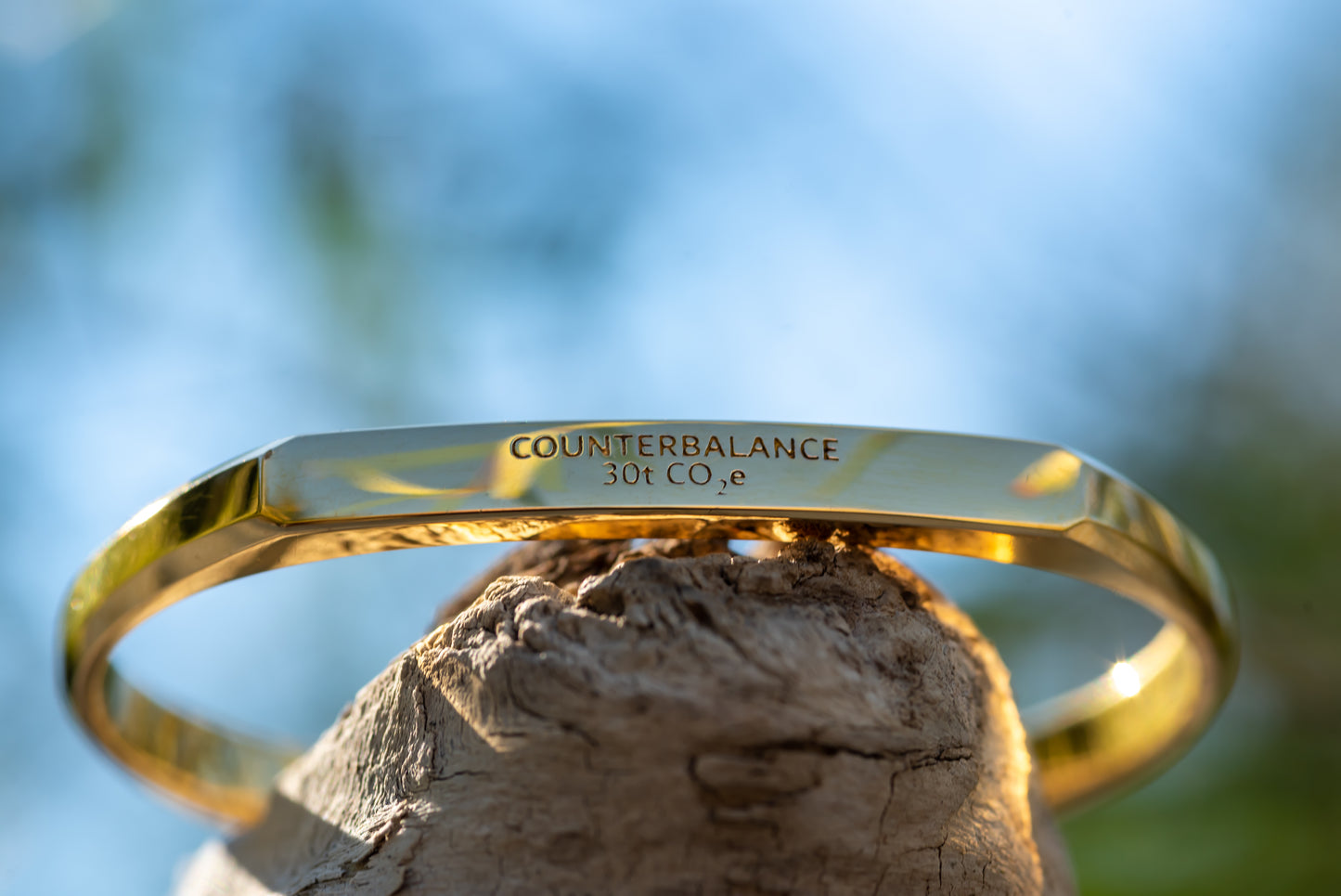 A gold counterbalance bracelet engraved with the abbreviations for 30 tons of carbon dioxide equivalent. The bracelet is displayed upon a piece of driftwood.