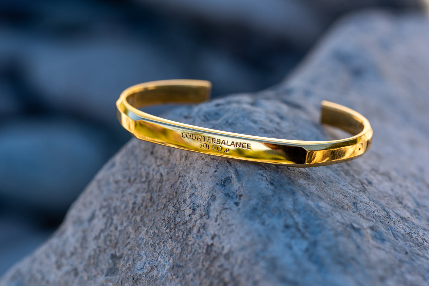 A gold counterbalance bracelet engraved with the abbreviations for 30 tons of carbon dioxide equivalent. The bracelet rests upon a grey rock.