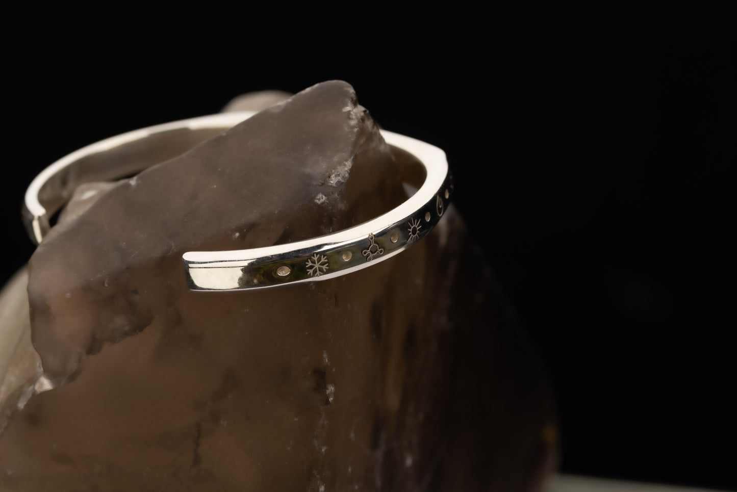 A silver counterbalance bracelet displayed on a piece of quartz engraved with several carbon offset climate action project charms. The charms from left to right are a snowflake for refrigerant management, a methane molecule for methane biofuels, a sun for solar energy, a flame for efficient cookstoves, and another sun for solar energy projects.