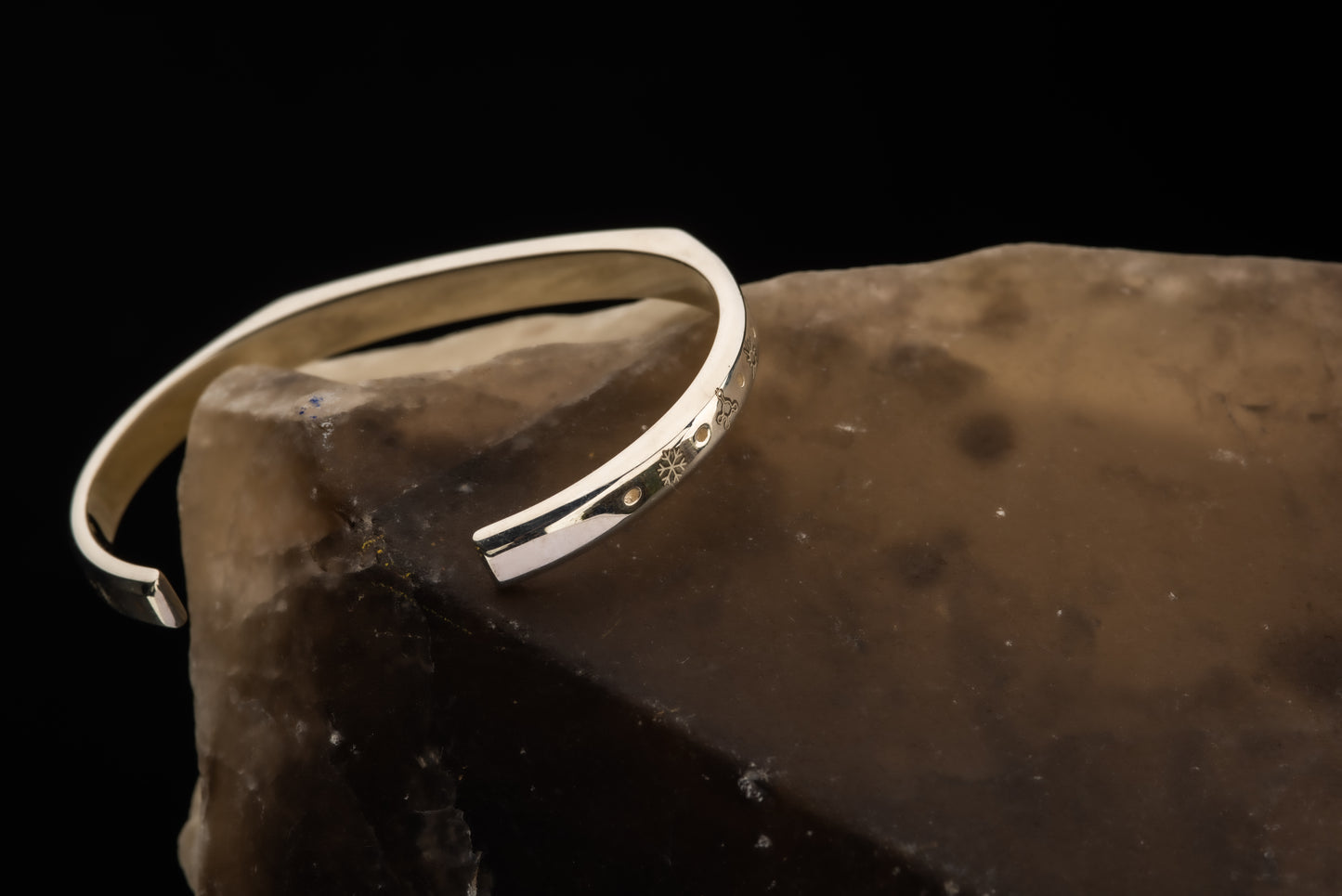 A silver counterbalance bracelet displayed on a piece of quartz engraved with several carbon offset climate action project charms. The charms from left to right are a snowflake for refrigerant management, a methane molecule for methane biofuels, a sun for solar energy, a flame for efficient cookstoves, and another sun for solar energy projects.