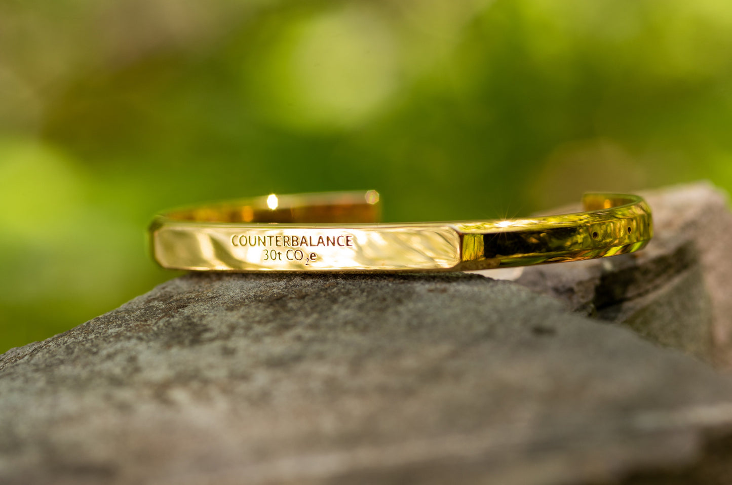 A gold counterbalance bracelet engraved with the abbreviations for 30 tons of carbon dioxide equivalent. The bracelet sits upon a lichen encrusted rock with a green forest background.