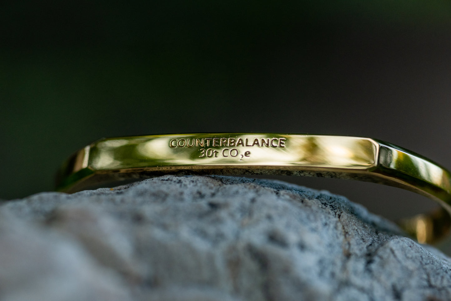 A gold counterbalance bracelet engraved with the abbreviations for 30 tons of carbon dioxide equivalent. The bracelet sits upon a smooth grey rock.