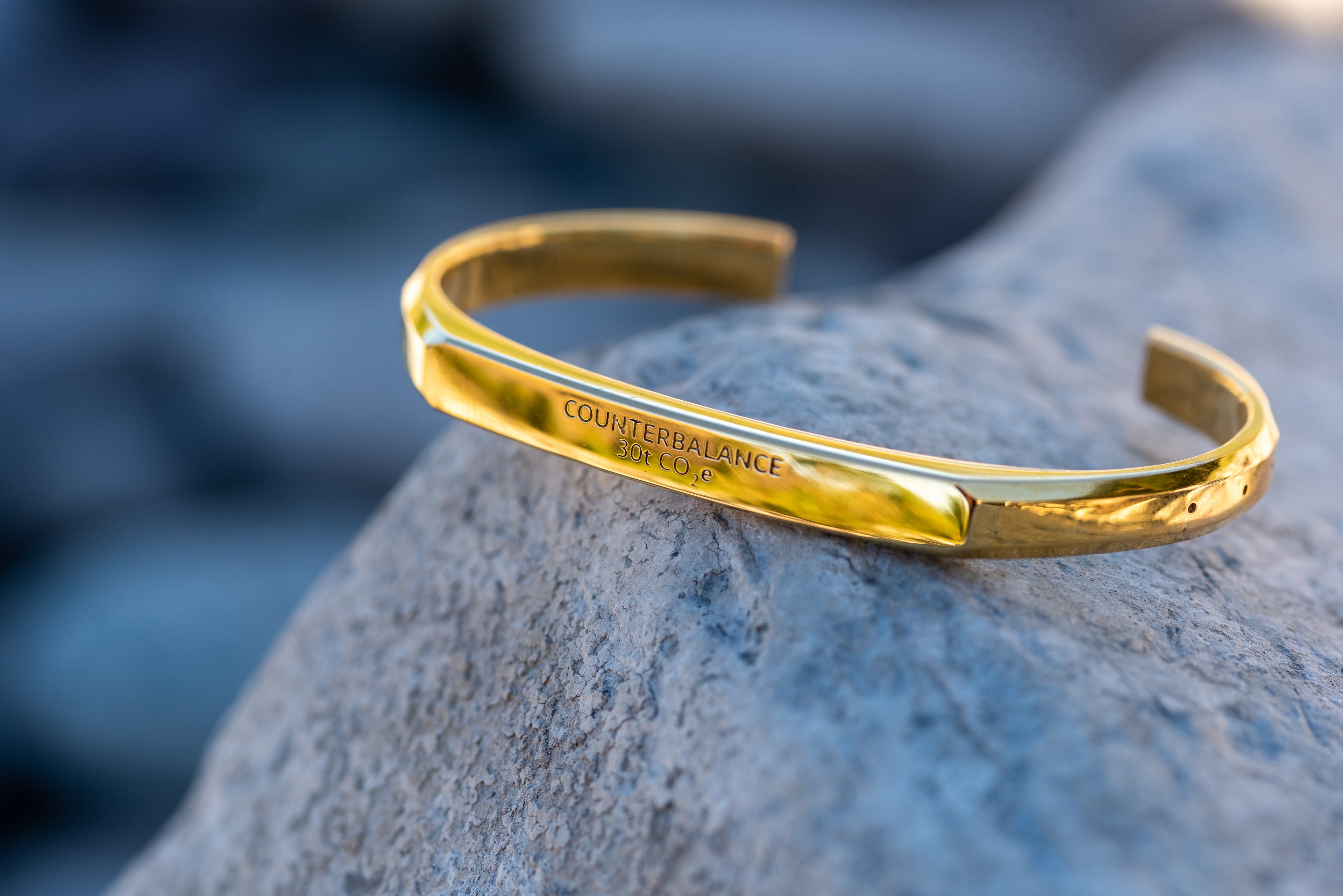 a gold counterbalance bracelet engraved with "30t CO2e" sits upon a grey blue rock. 