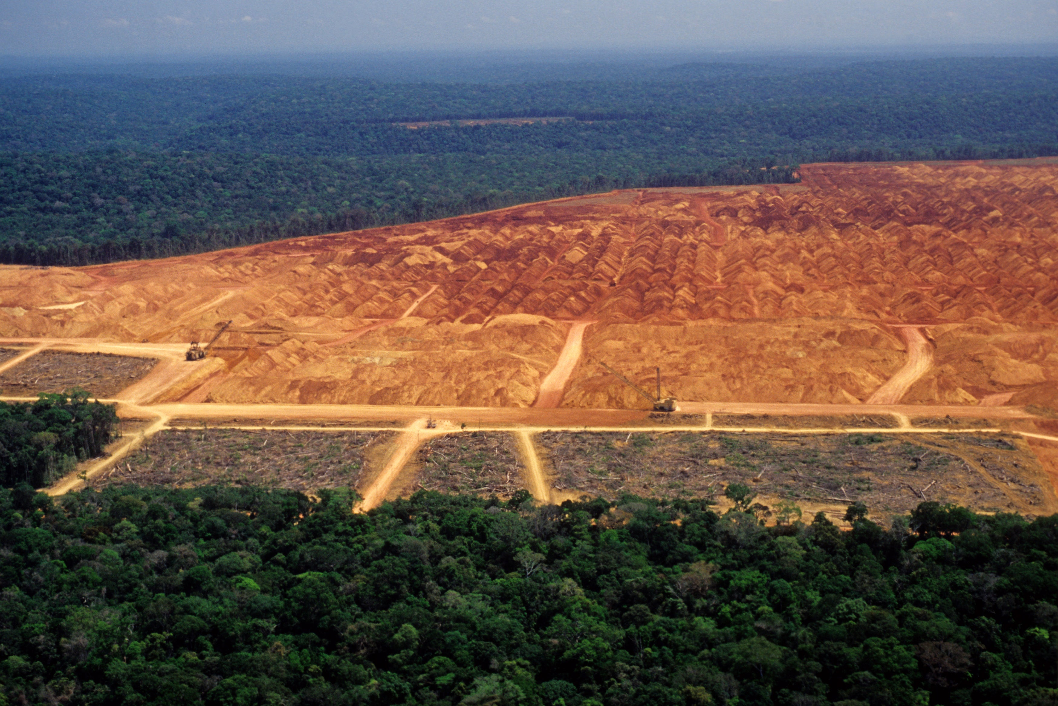 Deforestation in the amazon with red soil exposed with intact forest surrounding the area. Heavy machinery can be seen in the area. 