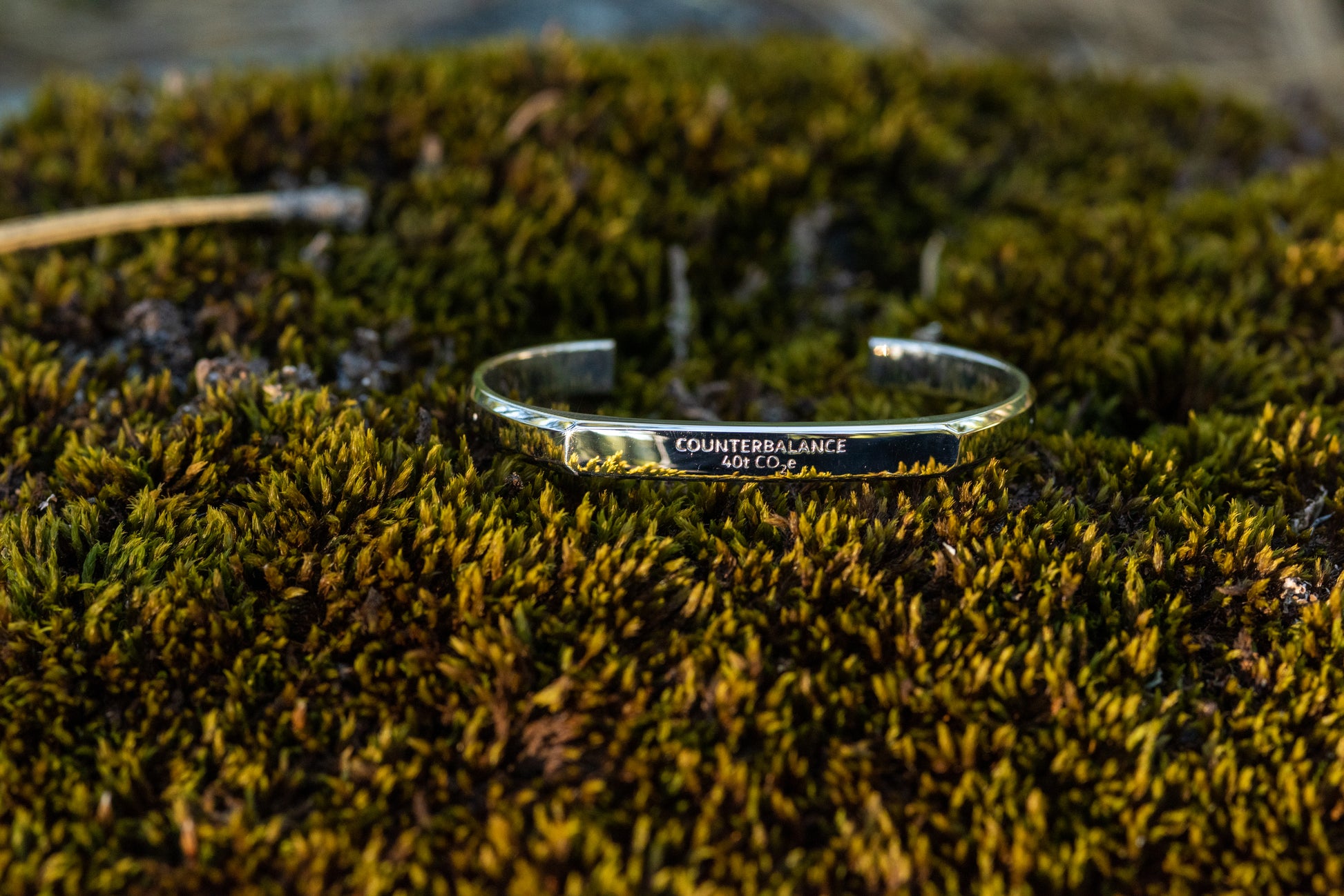 A white gold counterbalance bracelet engraved with 40t CO2e for 40 tons of carbon dioxide equivalent. The bracelet rests upon a mossy patch of forest floor. 