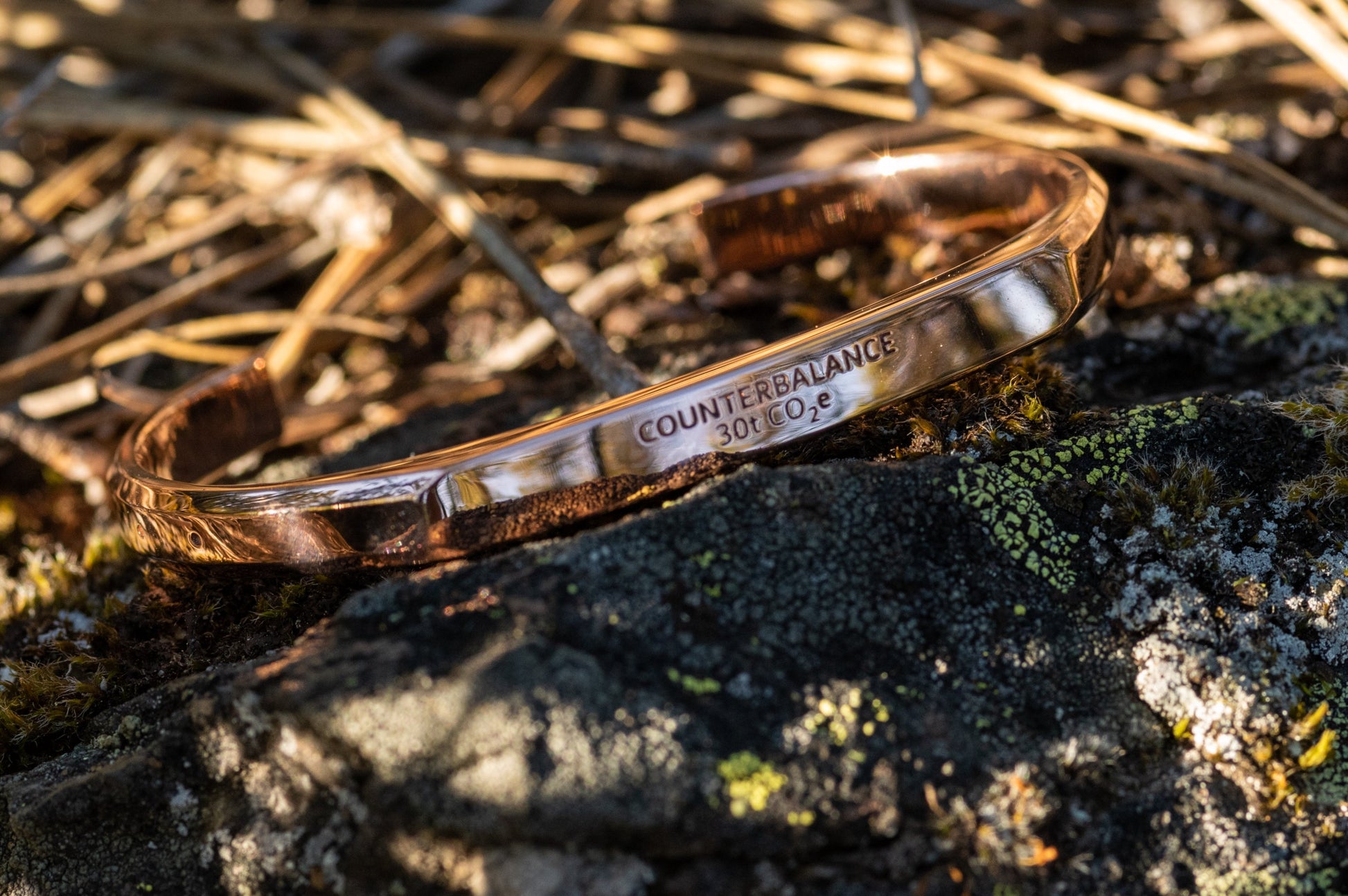 A rose gold counterbalance bracelet displayed upon piece mossy and lichen covered rock with ponderosa pine needles in the background. The bracelet is engraved with the word Counterbalance with the abbreviations and symbols for 30 tons of carbon dioxide equivalent below it.