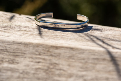 A white gold counterbalance bracelet engraved with 40t CO2e for 40 tons of carbon dioxide equivalent. The bracelet rests upon a piece of smooth driftwood with green sunlit foliage in the background.