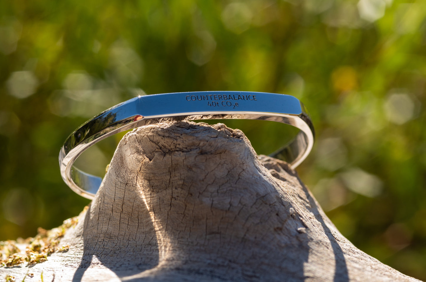 A white gold counterbalance bracelet engraved with 40t CO2e for 40 tons of carbon dioxide equivalent. The bracelet rests upon a piece of smooth driftwood with green sunlit foliage in the background.