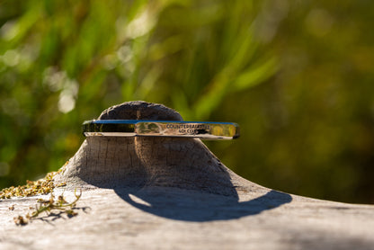 A white gold counterbalance bracelet engraved with 40t CO2e for 40 tons of carbon dioxide equivalent. The bracelet rests upon a piece of smooth driftwood with green sunlit foliage in the background.