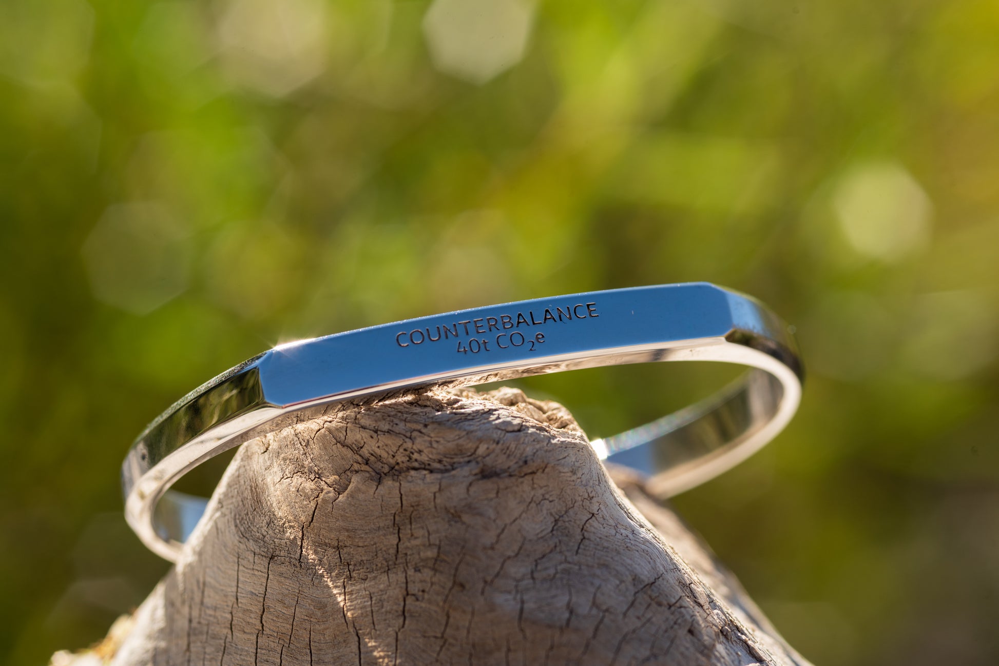 A white gold counterbalance bracelet engraved with 40t CO2e for 40 tons of carbon dioxide equivalent. The bracelet rests upon a piece of smooth driftwood with green sunlit foliage in the background. 