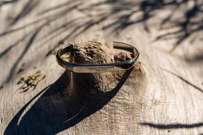 A white gold counterbalance bracelet engraved with 40t CO2e for 40 tons of carbon dioxide equivalent. The bracelet rests upon a piece of smooth driftwood with green sunlit foliage in the background.