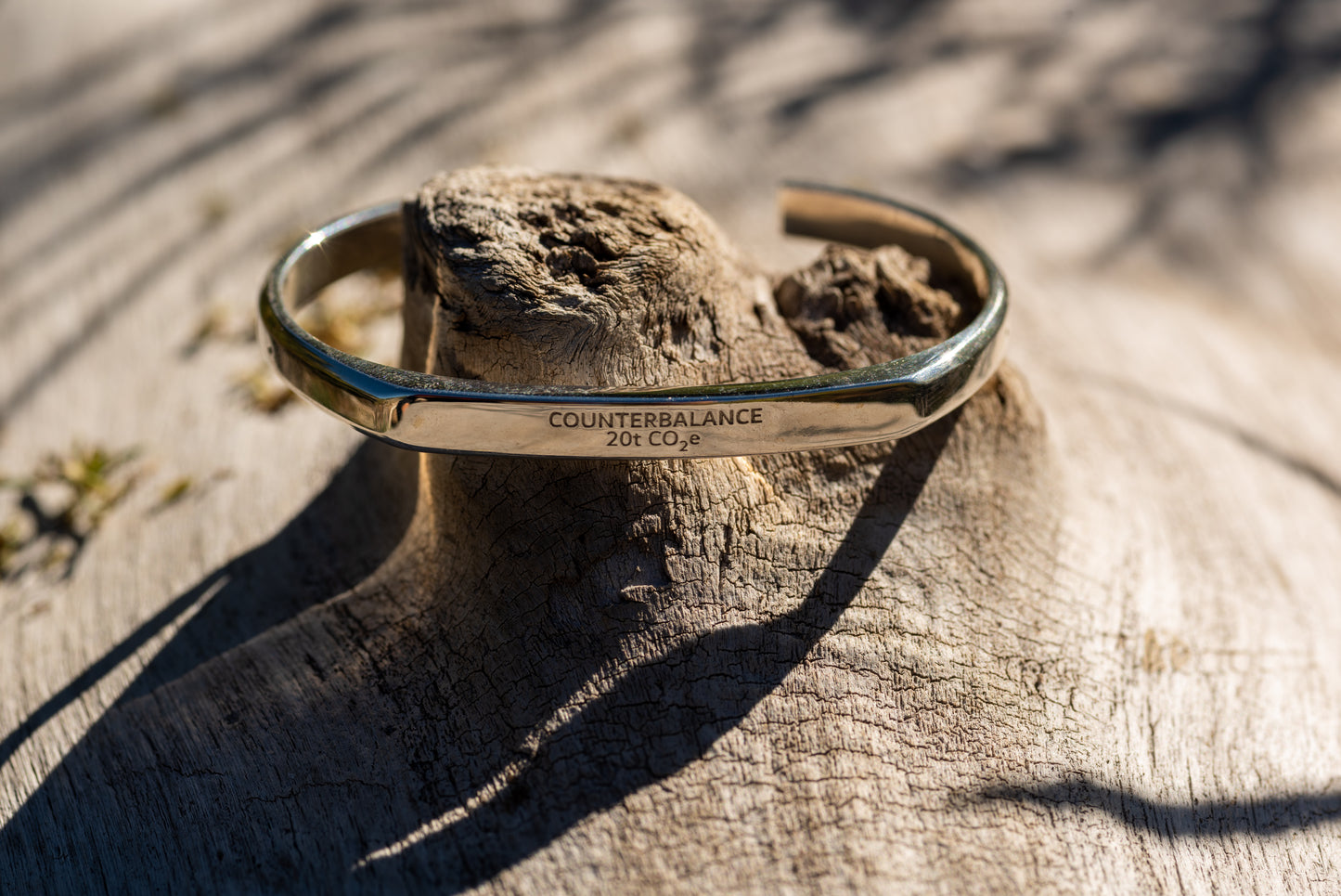 A silver counterbalance bracelet engraved with the word Counterbalance and the abbreviation 20t CO2e for 20 tons of carbon dioxide equivalent. The bracelet is displayed on a piece of smooth river driftwood. 