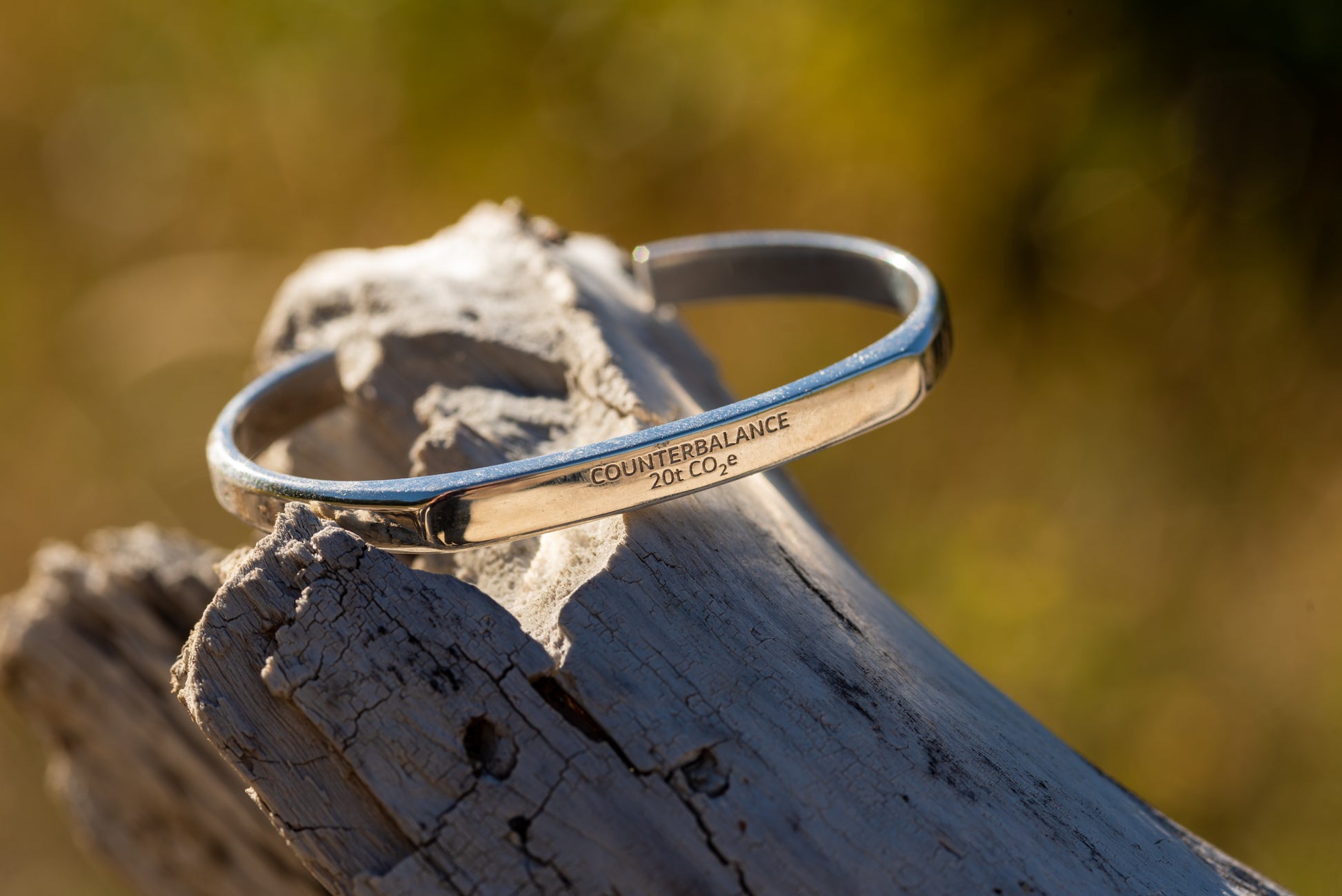 A silver counterbalance bracelet engraved with the word Counterbalance and the abbreviation 20t CO2e for 20 tons of carbon dioxide equivalent. The bracelet is displayed on a piece of driftwood.