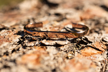 A rose gold counterbalance bracelet displayed upon ponderosa pine tree bark in a Montana forest. The bracelet is engraved with the word Counterbalance with the abbreviations and symbols for 30 tons of carbon dioxide equivalent below it.