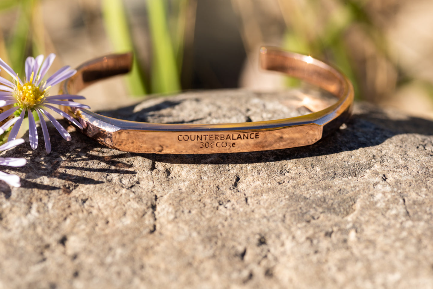 A rose gold counterbalance bracelet is on a rock adjacent to purple daisy wildflowers. The bracelet is engraved with the word Counterbalance with the abbreviations and symbols for 30 tons of carbon dioxide equivalent below it.