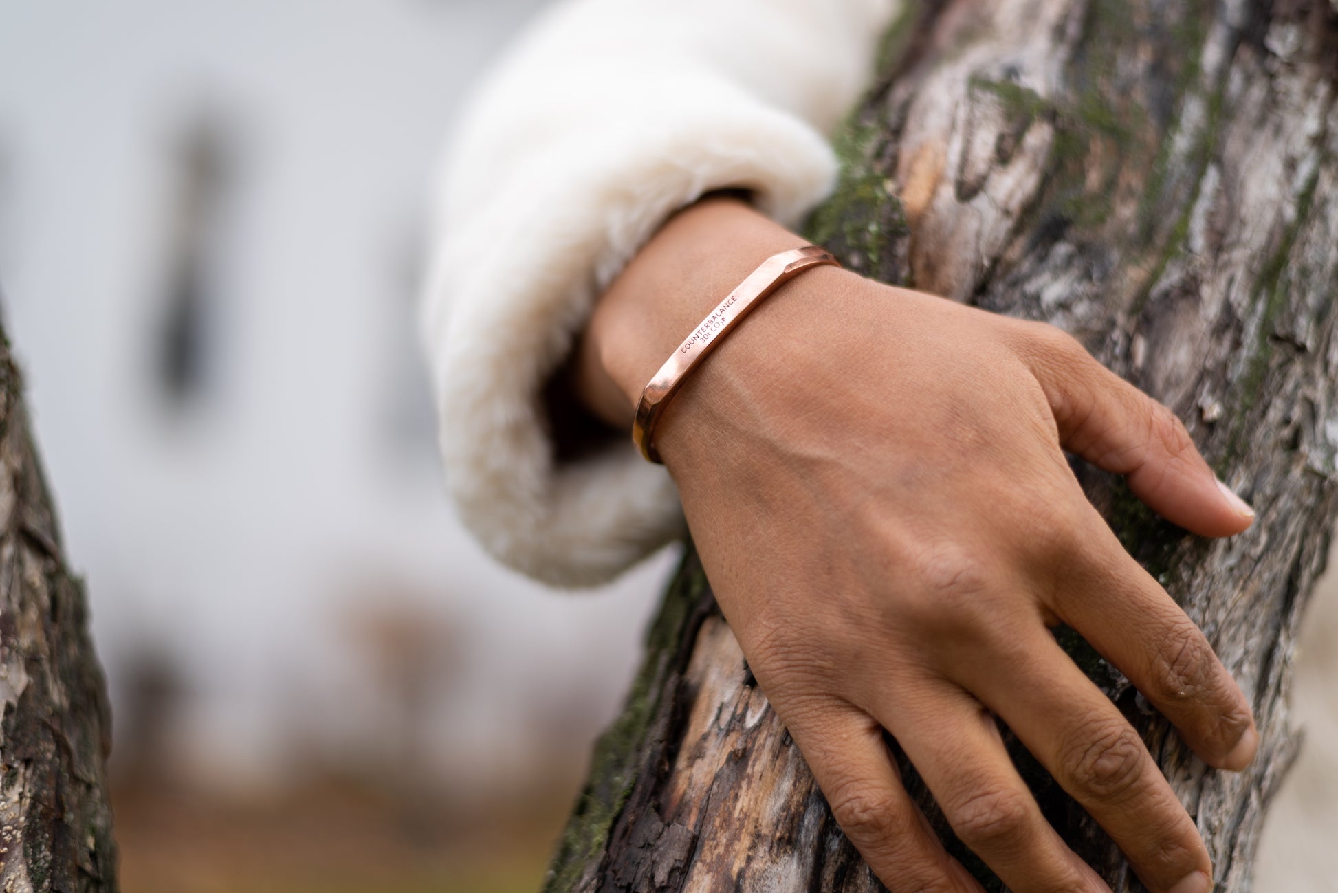 A rose gold counterbalance bracelet is worn by a model as she reaches around a tree trunk. The bracelet is engraved with the word Counterbalance with the abbreviations and symbols for 30 tons of carbon dioxide equivalent below it.