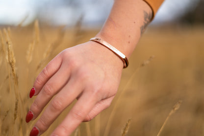 A rose gold counterbalance bracelet is worn by a model in tall golden grasses. The bracelet is engraved with the word Counterbalance with the abbreviations and symbols for 30 tons of carbon dioxide equivalent below it.