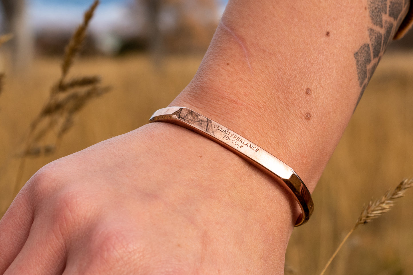 A rose gold counterbalance bracelet is worn by a model in tall golden grasses. The bracelet is engraved with the word Counterbalance with the abbreviations and symbols for 30 tons of carbon dioxide equivalent below it.