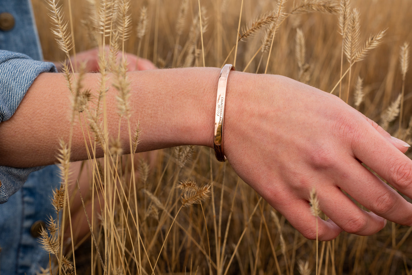 A rose gold counterbalance bracelet is worn by a model in tall golden grasses. The bracelet is engraved with the word Counterbalance with the abbreviations and symbols for 30 tons of carbon dioxide equivalent below it.