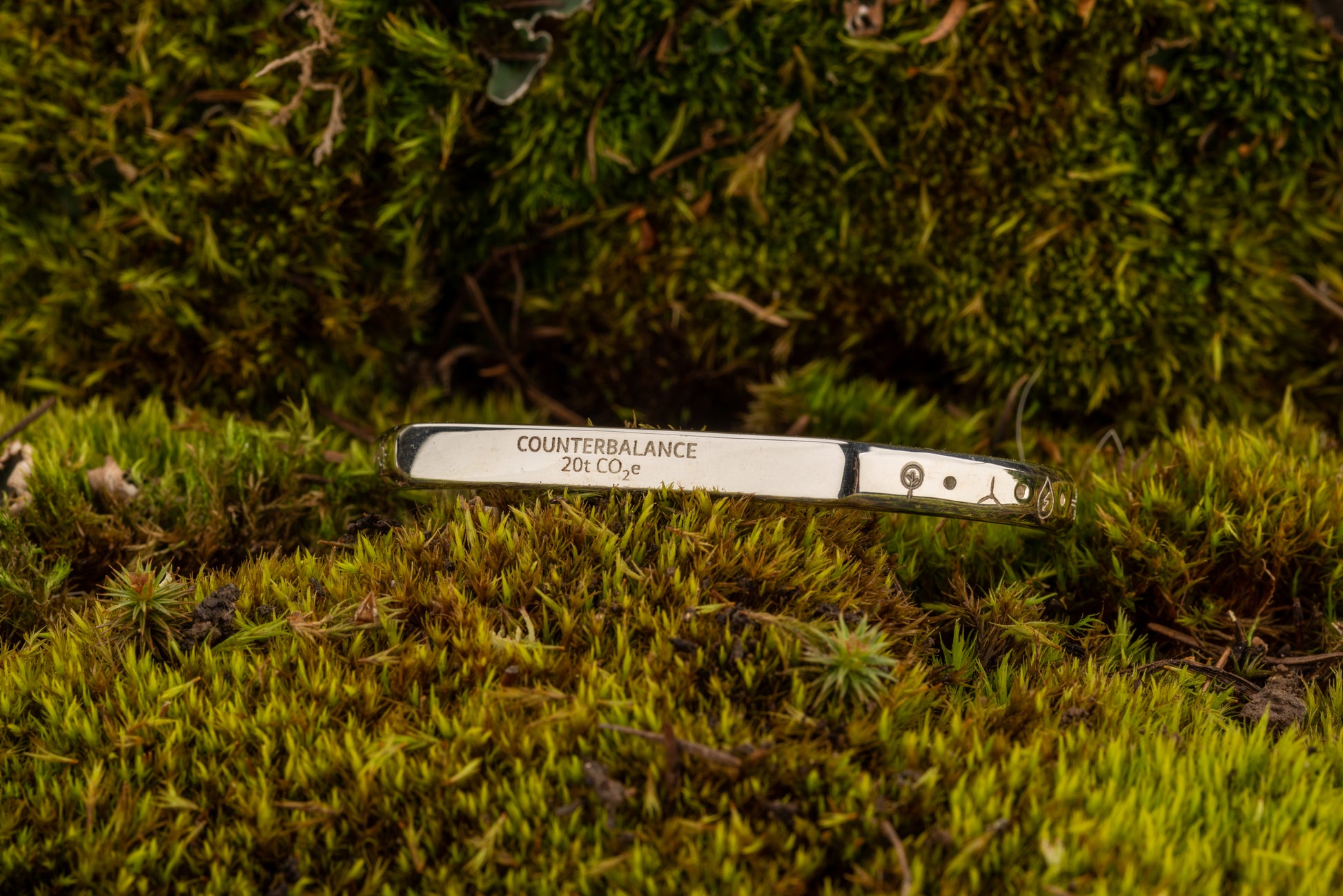 A silver counterbalance bracelet engraved with the word Counterbalance and the abbreviation 20t CO2e for 20 tons of carbon dioxide equivalent. The bracelet is displayed on a bed of moss.