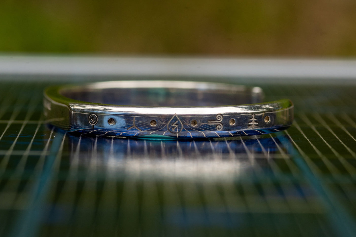 A silver counterbalance bracelet displayed on a solar panel. The bracelet is engraved with several carbon offset climate action project charms. The charms from left to right display a simple tree icon for forest conservation, a wind turbine for wind energy , a water droplet for hydroelectric energy, a wind symbol for premium wind projects, and an evergreen tree for forest conservation and restoration projects.