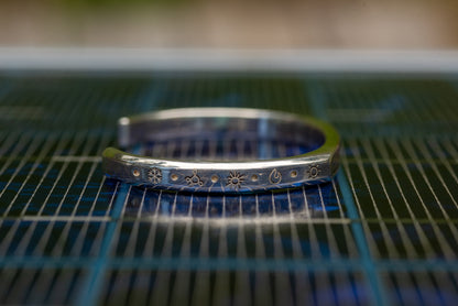 A silver counterbalance bracelet displayed on a solar panel. The bracelet is engraved with several carbon offset climate action project charms. The charms from left to right are a snowflake for refrigerant management, a methane molecule for methane biofuels, a sun for solar energy, a flame for efficient cookstoves, and another sun for solar energy projects.