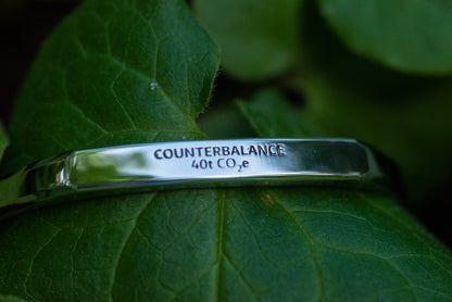 A white gold counterbalance bracelet engraved with 40t CO2e for 40 tons of carbon dioxide equivalent. The bracelet rests upon a soft green leaf.